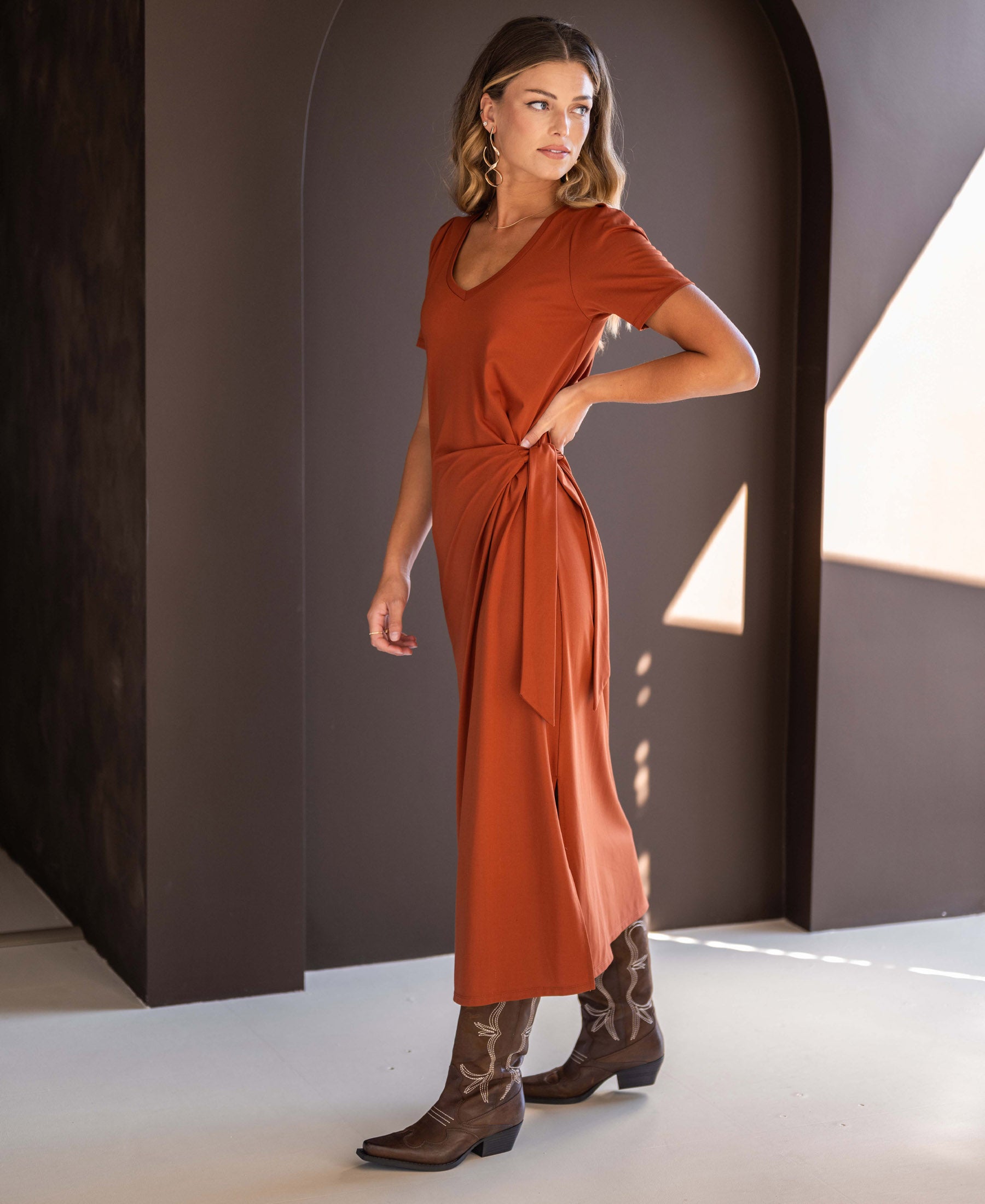 Woman wears the PLEIN PUBLIQUE long dress LA FILICIA in terra color, standing by a dark arched wall.