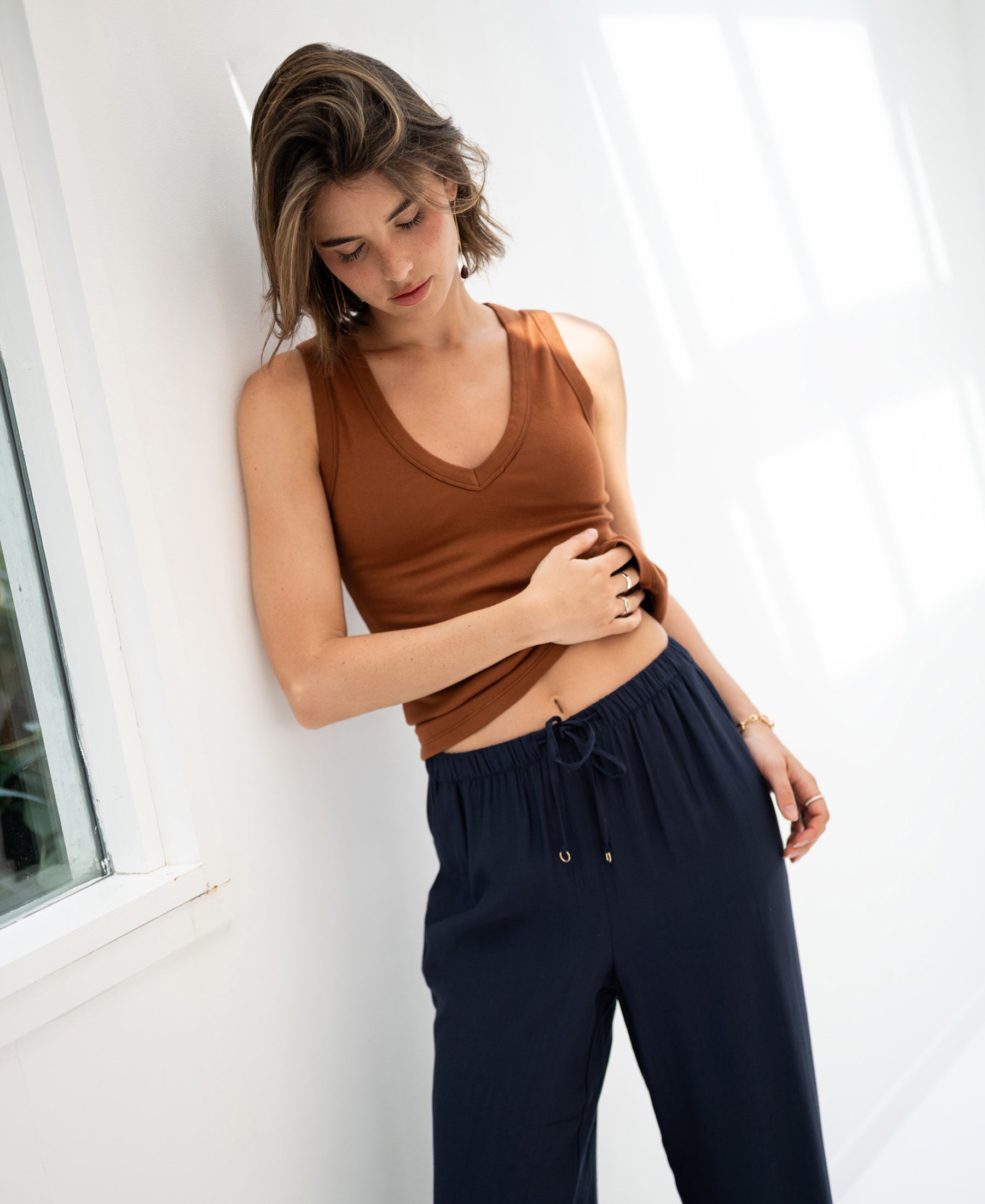 Woman wears a luxe and soft brown LA VALE top by PLEIN PUBLIQUE, leaning against a white wall and looking down.