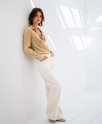 Woman wears the satin stretch blouse LE VENT in light sand by PLEIN PUBLIQUE, hands in pockets.