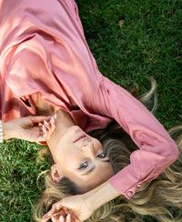 Dusty pink satin stretch blouse displayed outdoors, showing front view as worn by a model lying on grass.