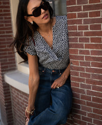 Woman wears charcoal and ivory dotted Stippen top by PLEIN PUBLIQUE, leaning on a brick wall.