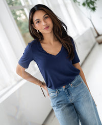 Woman wears a bamboo T-shirt in denim blue, standing indoors with a plant and windows in the background.