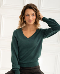 Green merino wool V-neck sweater, shown indoors on a model with wavy hair, hand resting on head.