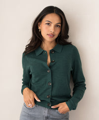 Woman wearing a greenish collared vest, standing by a light wall.