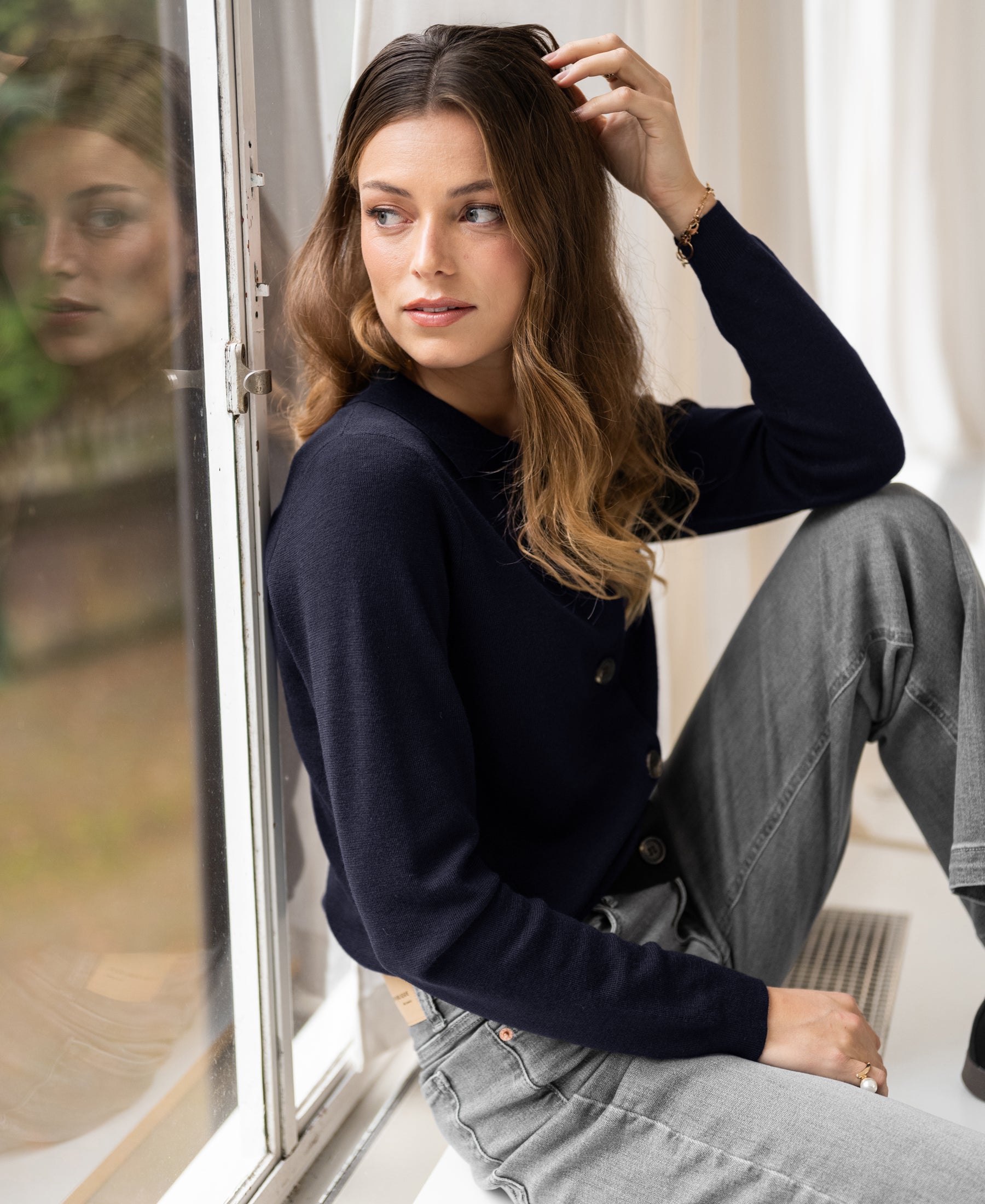Dark blue LES VINGT polo-collar cardigan by PLEIN PUBLIQUE, shown seated by a window with reflection visible.