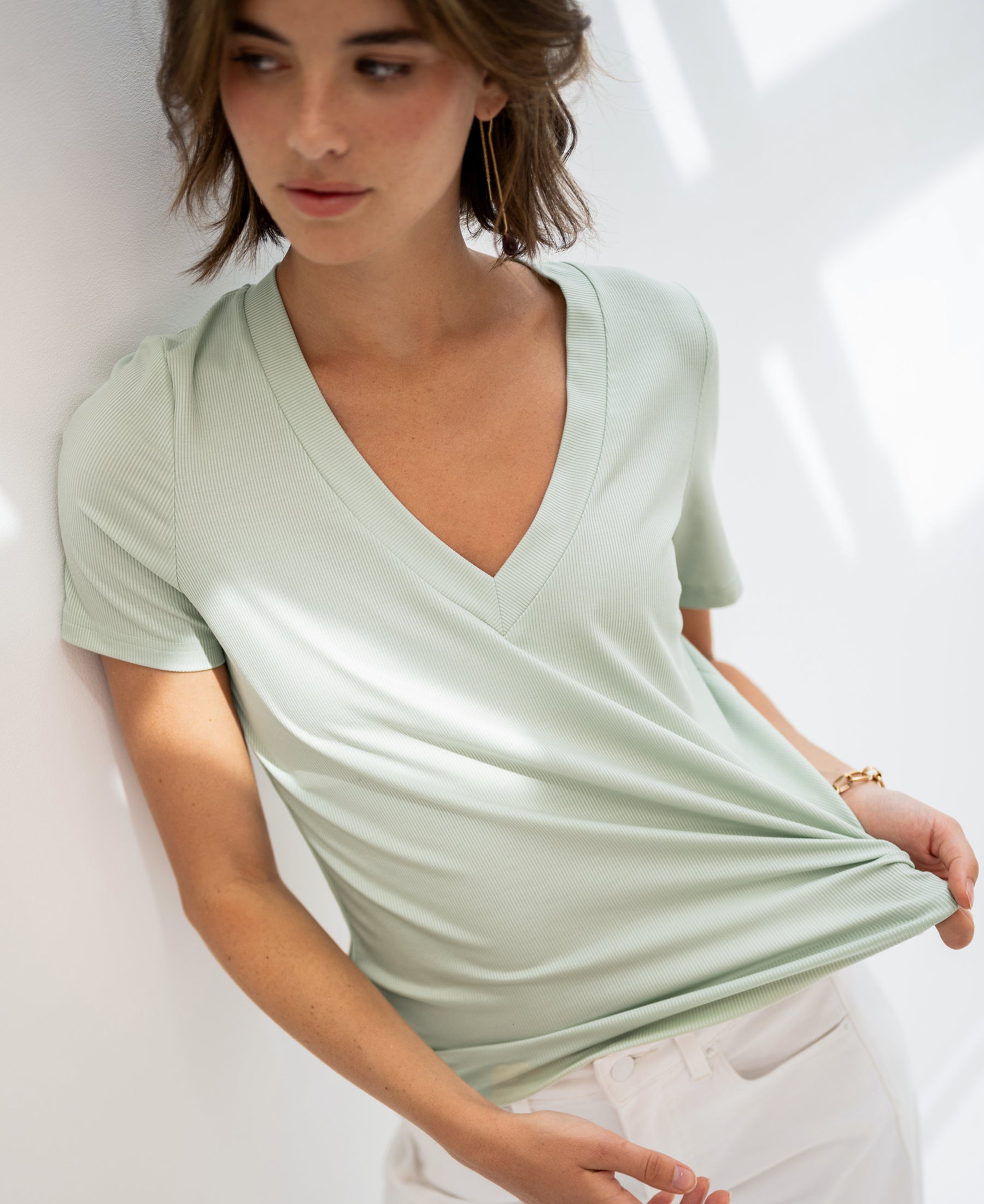 Woman wearing a light green lyocell T-shirt stands by a white wall in soft sunlight.