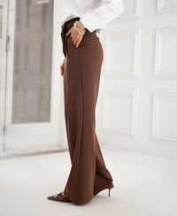 Woman wearing brown PLEIN PUBLIQUE LA WENDE pants in a minimal, bright white-walled room.