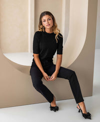 Woman wears black merino wool sweater, seated on a beige arch in a minimalist space.