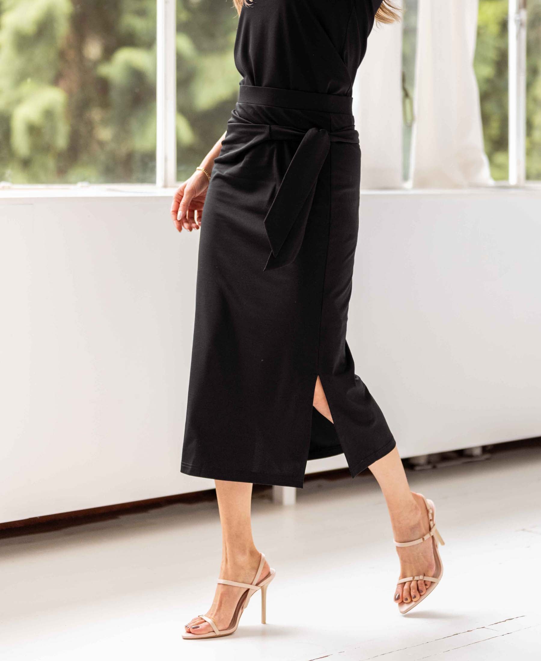 Woman wearing a black LE MAEL Travel skirt by PLEIN PUBLIQUE, walking indoors near windows with greenery visible outside.