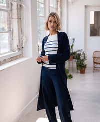 Woman wearing a PLEIN PUBLIQUE Breton sweater LELISA in ivory and denim blue stands by plants and large windows.
