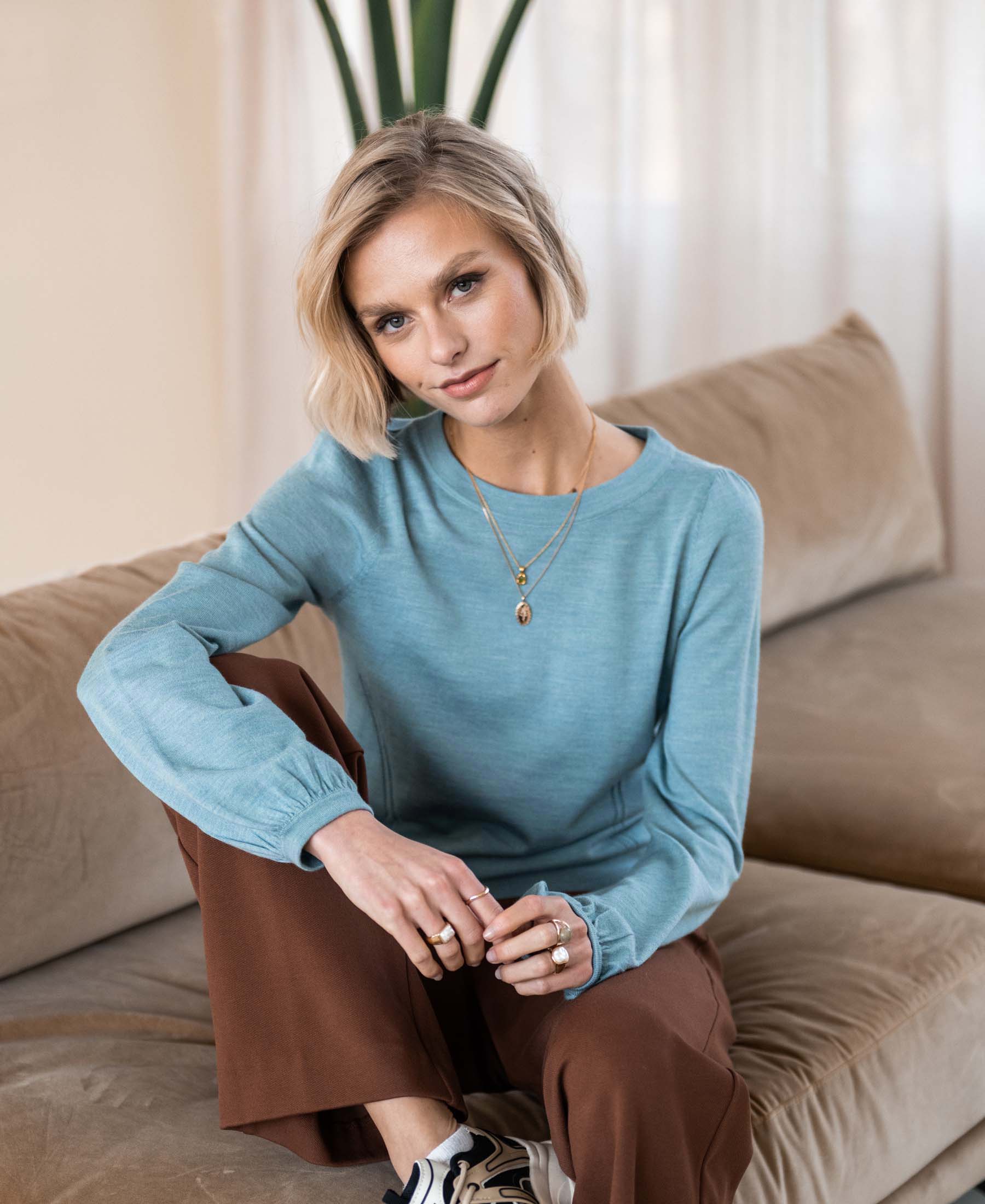 Mist Blue LA COEUR merino wool sweater from PLEIN PUBLIQUE, shown on a model sitting on a beige sofa.