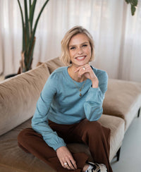 Merino wool LA COEUR sweater in light blue by PLEIN PUBLIQUE, shown worn while seated on a beige couch.