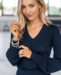 A woman with long blonde hair and blue eyes, wearing the Merino V-hals trui LA VICTORIA Donkerblauw, hoop earrings, bracelets, and rings, stands indoors and smiles slightly at the camera.