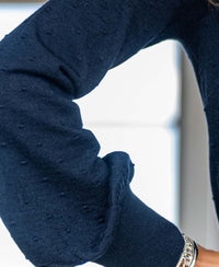 Close-up of a person's bent arm wearing the Merino V-hals sweater LA VICTORIA Donkerblauw with textured navy knit and a silver chain bracelet, set against a light background.