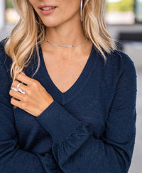 Woman with long blonde hair wearing the Merino V-hals sweater LA VICTORIA Donkerblauw, styled with a silver necklace and rings. Her face is partially visible, hand in her hair, against a blurred background—ideal for elegante dames knitwear fans.