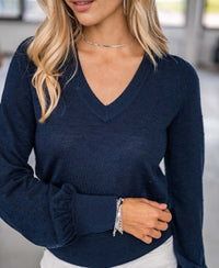 A woman with long blonde hair wears the Merino V-hals trui LA VICTORIA Donkerblauw, paired with white pants, a silver necklace, and bracelets. She stands indoors with her hand on her waist.