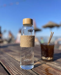 The glass water bottle with bamboo cap by PLEIN PUBLIQUE lies on a wooden table.