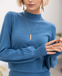 Woman wearing the PLEIN PUBLIQUE turtleneck sweater in steel blue, hands clasped, facing forward.