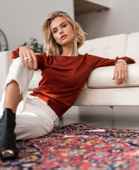 Woman wearing PLEIN PUBLIQUE Merino sweater LA COEUR in rosewood, seated on a colorful rug near a white sofa.