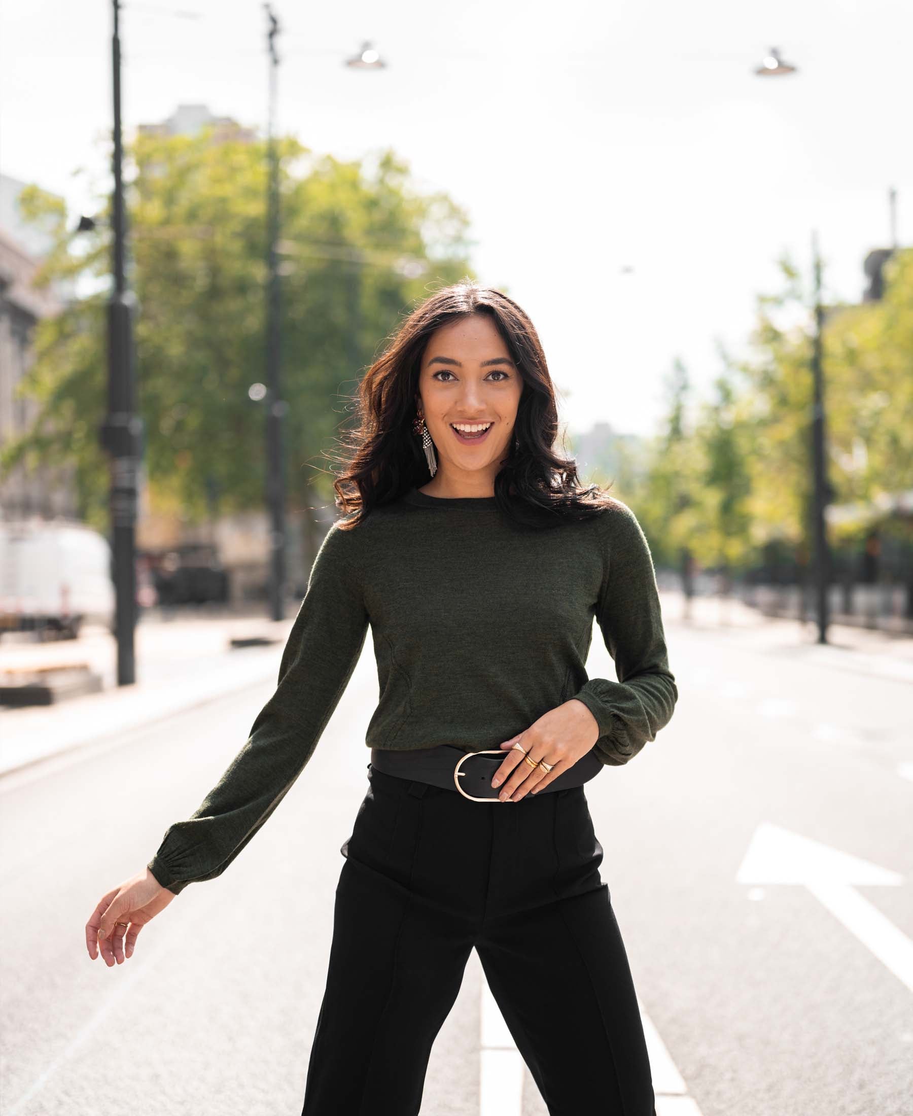 Woman wearing a green Merino wool LA COEUR sweater by PLEIN PUBLIQUE stands on a sunlit street.