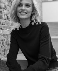 Woman indoors wearing a navy blue merino wool sweater, shown in front of a brick wall.