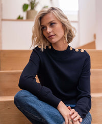 Woman wearing a PLEIN PUBLIQUE dark blue merino wool sweater sits on wooden stairs.