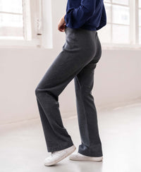 Woman wearing PLEIN PUBLIQUE knitted trousers LE LOURDES in anthracite stands by large windows indoors.