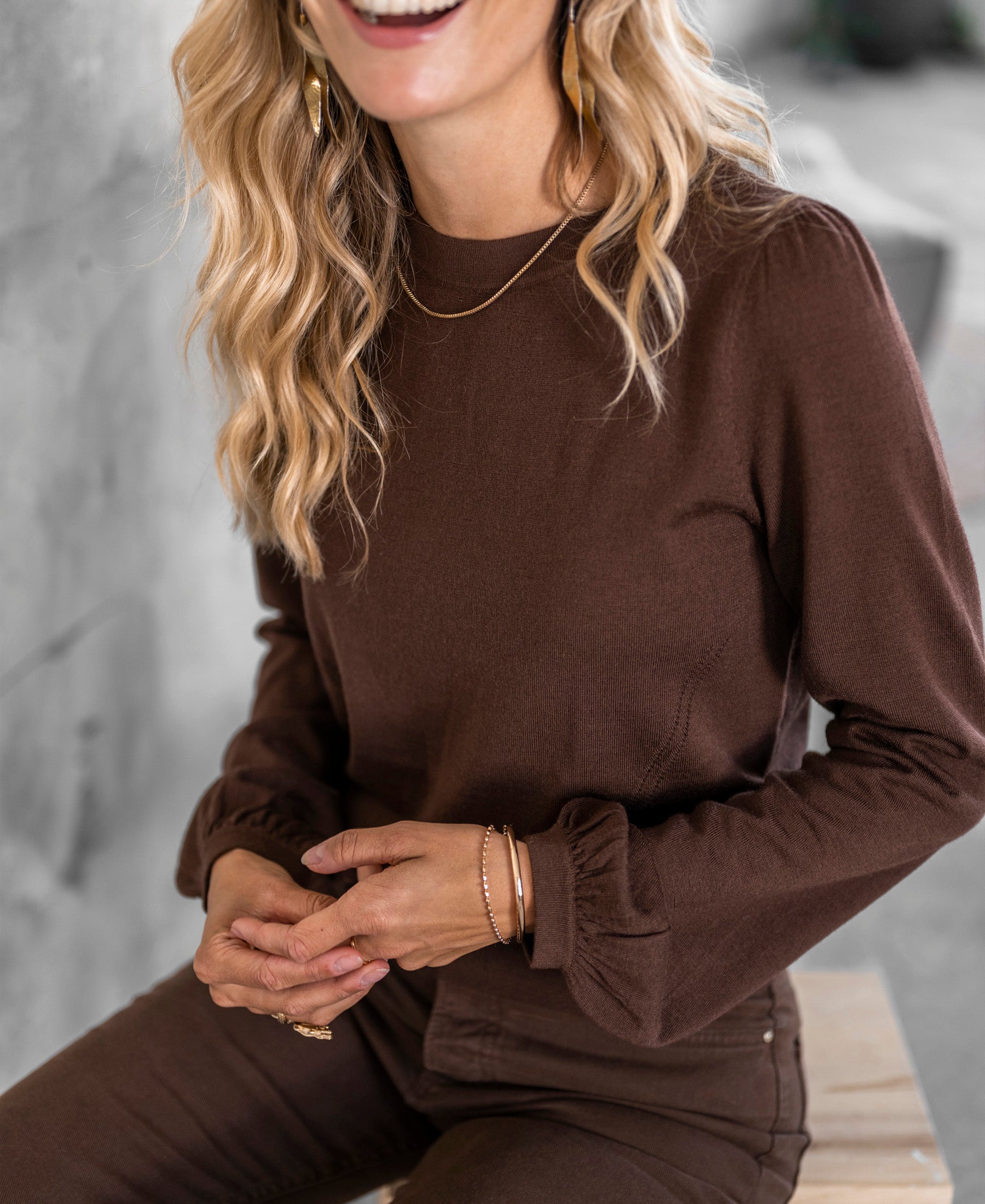 Brown merino wool sweater, front view, shown on a model sitting on a wooden bench.