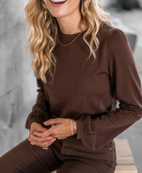 Brown merino wool sweater, front view, shown on a model sitting on a wooden bench.