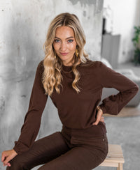 Woman wearing the brown LA COEUR Merino sweater by PLEIN PUBLIQUE, shown in a modern interior.
